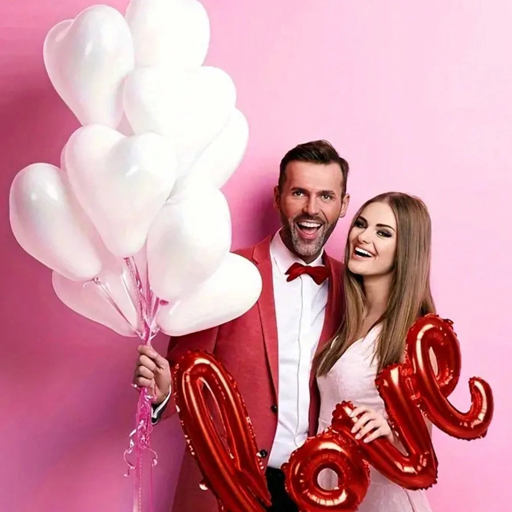 A man and a woman smiling while holding a bouquet of white heart-shaped latex balloons and red foil letters.