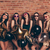 Five young women posing with black and gold latex balloons against a brick wall.