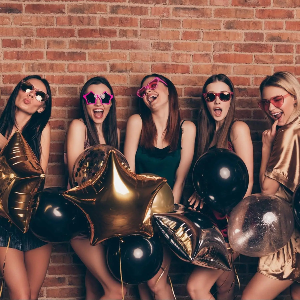 Five young women posing with black and gold latex balloons against a brick wall.