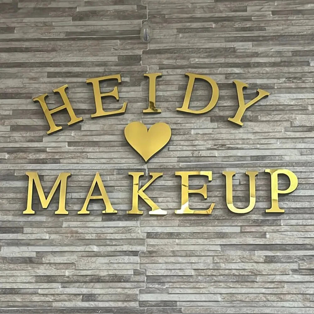 Gold acrylic mirror letter wall stickers spelling HEIDY MAKEUP with a heart accent on a gray stone background