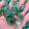 A hand holds several raw green and blue-green crystal stones on a textured surface.