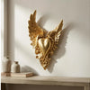 Gold ornate winged heart wall decor mounted on a white wall above a wooden shelf.
