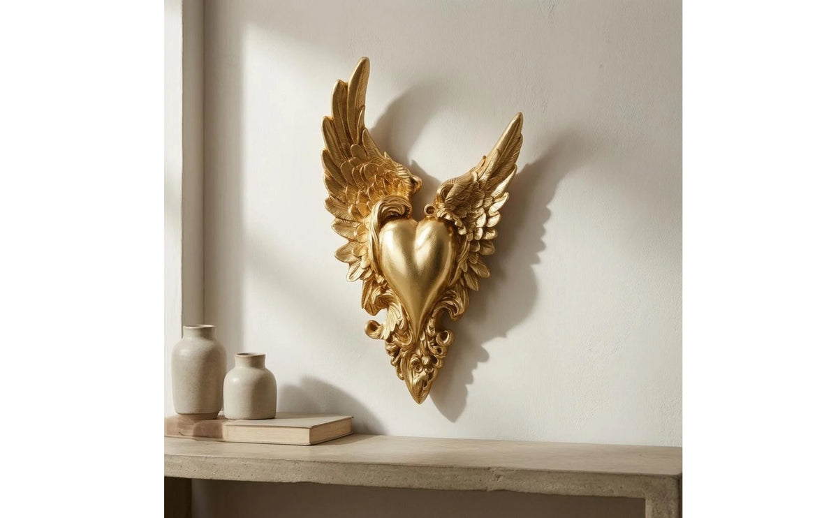 Gold ornate winged heart wall decor mounted on a white wall above a wooden shelf.