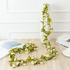 A silk rose garland with green leaves and white flowers is displayed on a wooden surface next to a white vase.