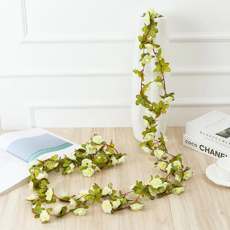 A silk rose garland with green leaves and white flowers is displayed on a wooden surface next to a white vase.