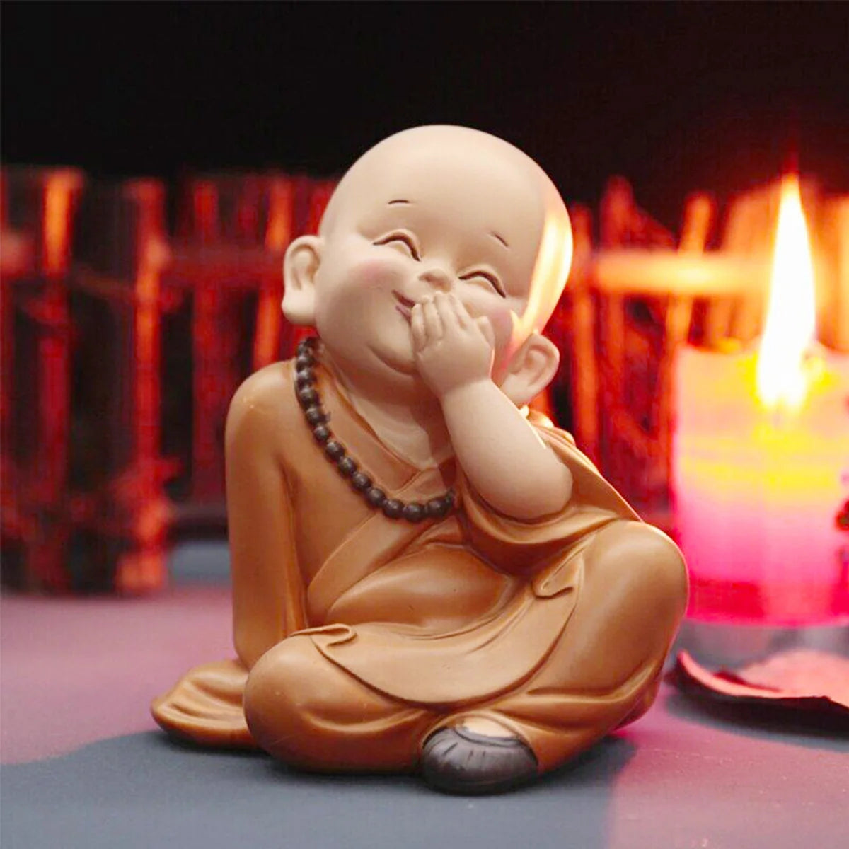 A small, light brown resin figurine of a smiling monk with hands clasped, sitting on a surface next to a glowing candle.