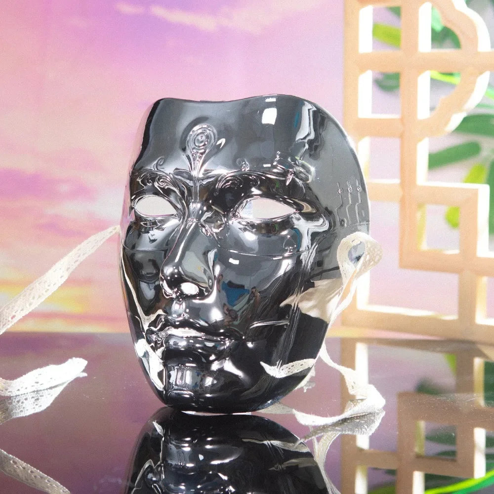 A glossy black ancient style masquerade mask with intricate facial details is displayed on a reflective surface.