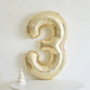 A large champagne gold foil number balloon shaped like the number three is displayed on a white surface.