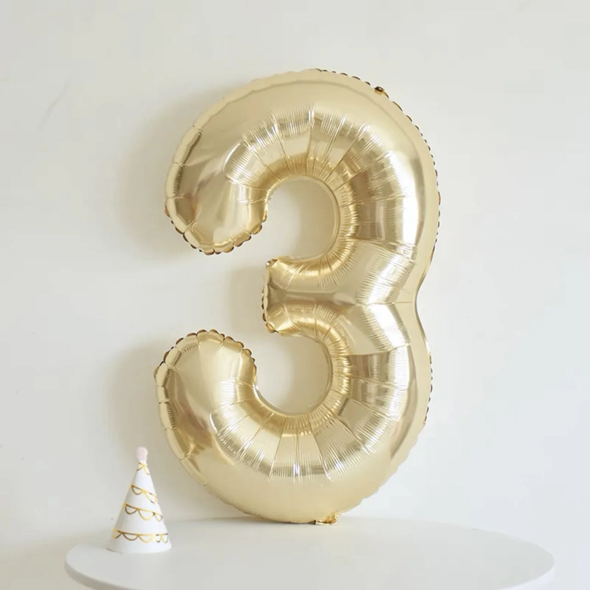 A large champagne gold foil number balloon shaped like the number three is displayed on a white surface.