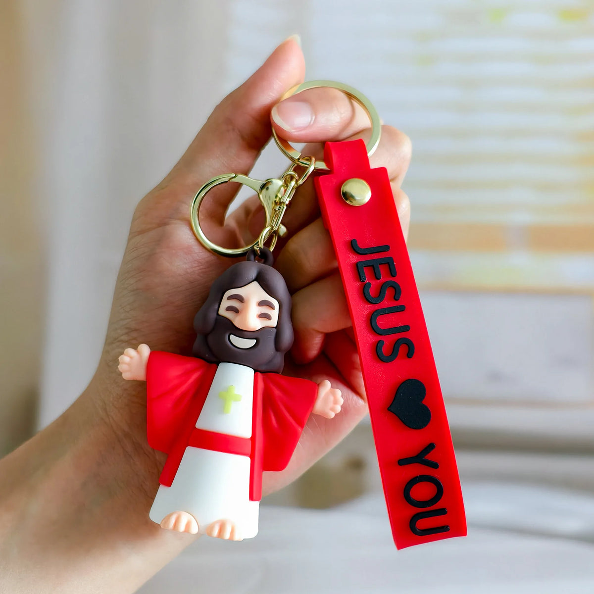 A hand holds a silicone Jesus keychain with a red strap and gold split ring.