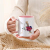 A person holds a white ceramic coffee mug with a gold letter initial decorated with colorful flowers.