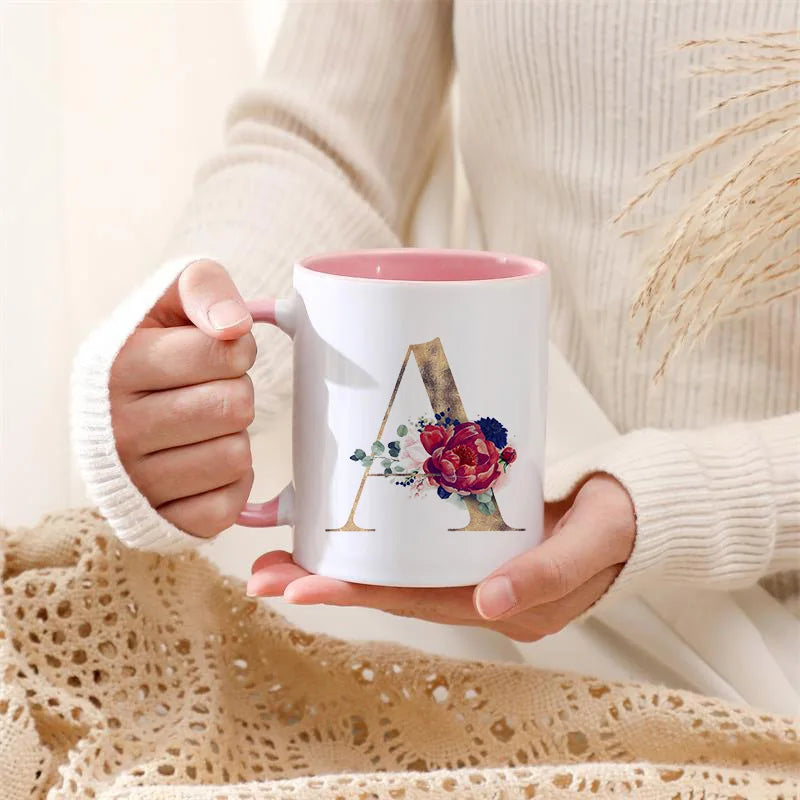 A person holds a white ceramic coffee mug with a gold letter initial decorated with colorful flowers.