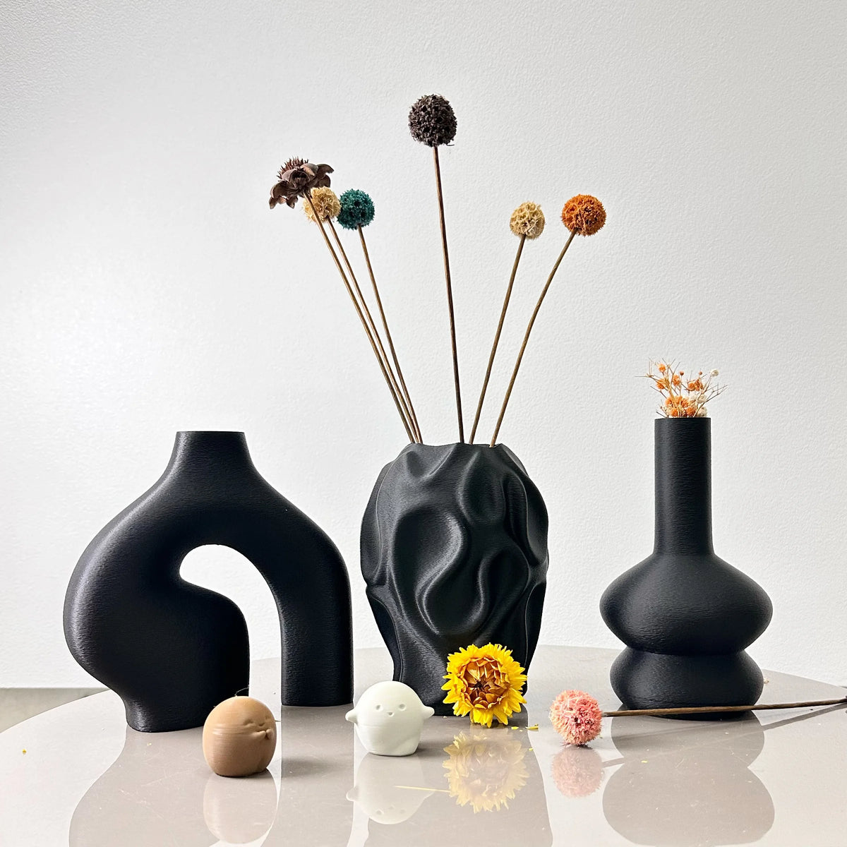 A black geometric vase set featuring three dried floral stems with spherical seed heads on a white surface.