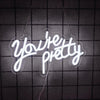 A white neon sign spelling out You're pretty is suspended against a dark grid background.
