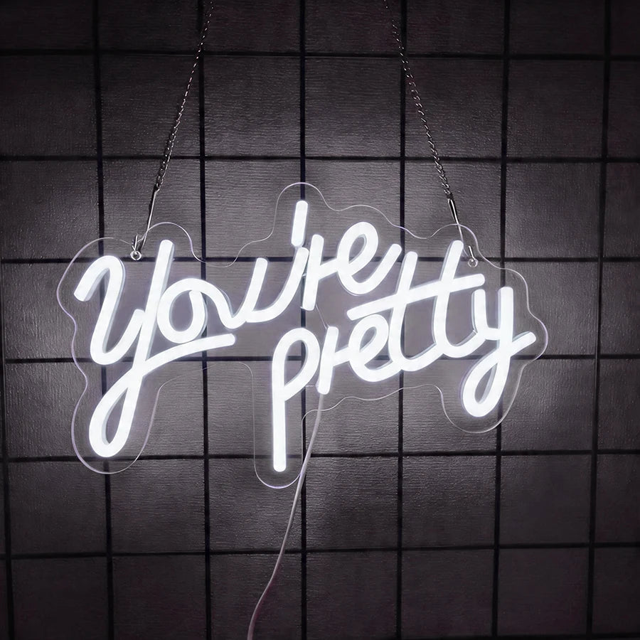 A white neon sign spelling out You're pretty is suspended against a dark grid background.