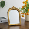 A vintage arched carved gold vanity mirror with a reflective surface sits on a dark wooden table.