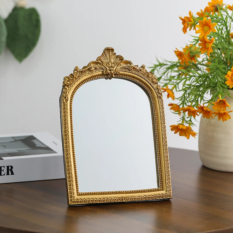 A vintage arched carved gold vanity mirror with a reflective surface sits on a dark wooden table.