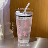 A clear plastic cup with a pink bow design, straw, and lid, shown against a wooden background.