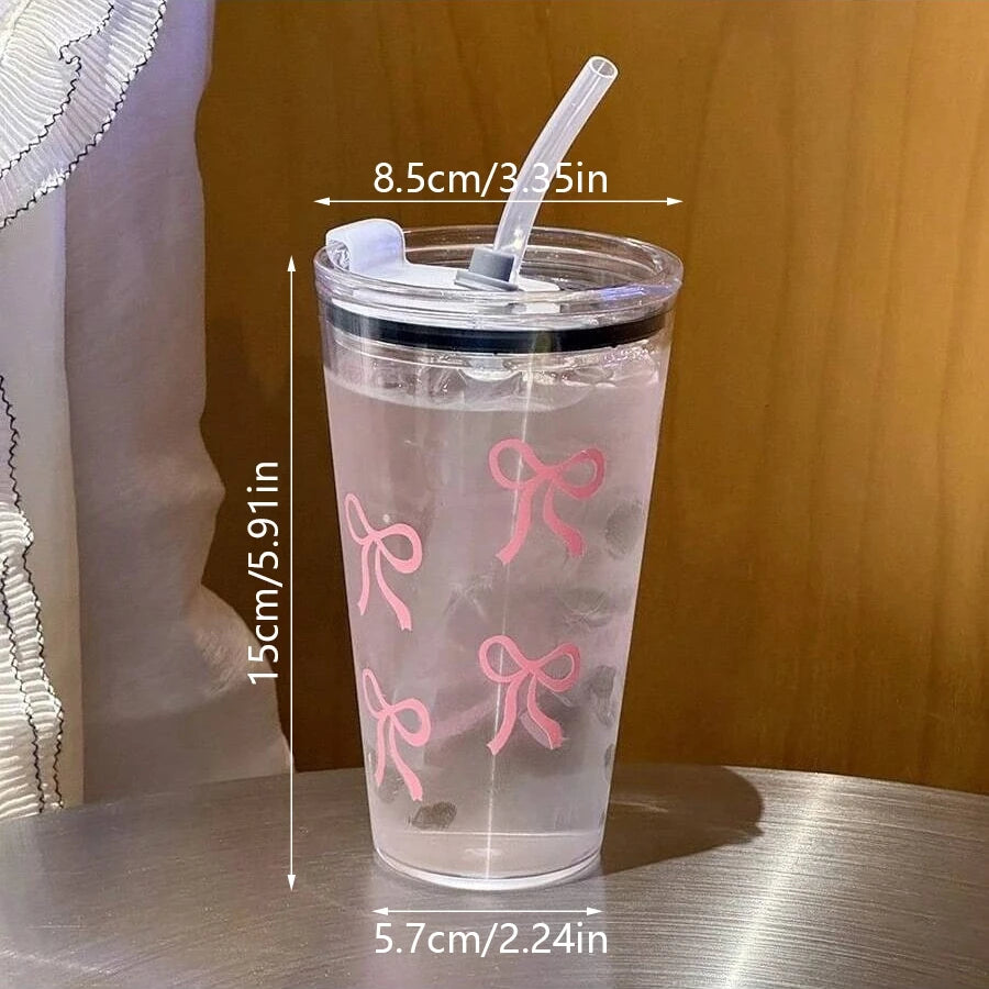 A clear plastic cup with a pink bow design, straw, and lid, shown against a wooden background.