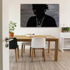 A dark portrait of a person wearing a necklace is displayed on a canvas above a wooden dining table.