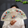 A person holds an open hand-painted decorative folding fan with floral designs against a dark background.