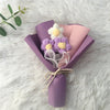 A handcrafted crochet sunflower bouquet featuring purple and white flowers wrapped in lilac paper on a white fluffy surface.