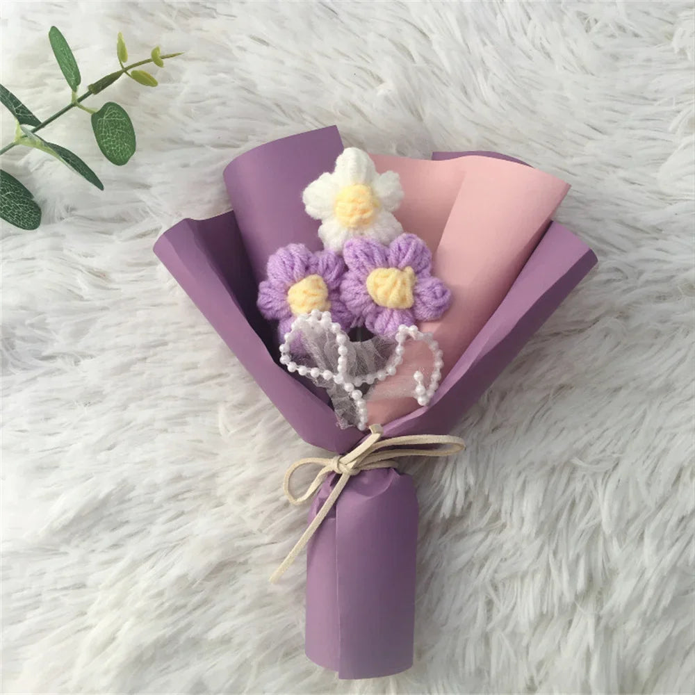 A handcrafted crochet sunflower bouquet featuring purple and white flowers wrapped in lilac paper on a white fluffy surface.