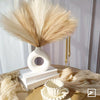 A white vase holds dried pampas grass decor alongside gold decorative sticks on a wooden surface.