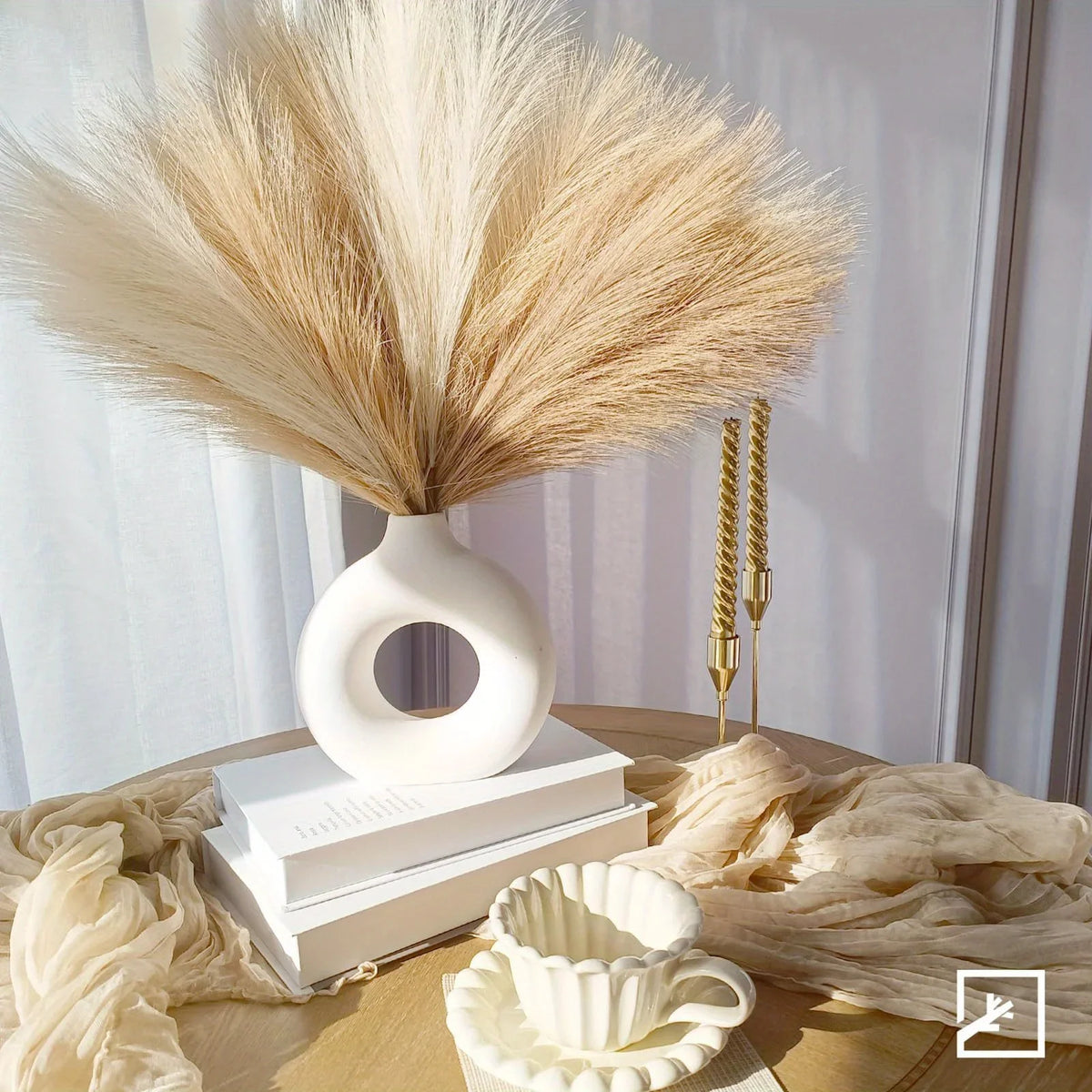 A white vase holds dried pampas grass decor alongside gold decorative sticks on a wooden surface.