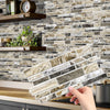 A hand holds a rectangular panel of crystal film marble wall decal against a textured stone tile background.