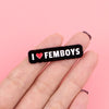 A hand holds a black enamel pin with the text I love femboys engraved on it.