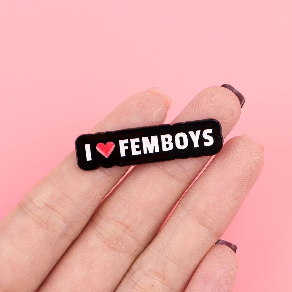 A hand holds a black enamel pin with the text I love femboys engraved on it.