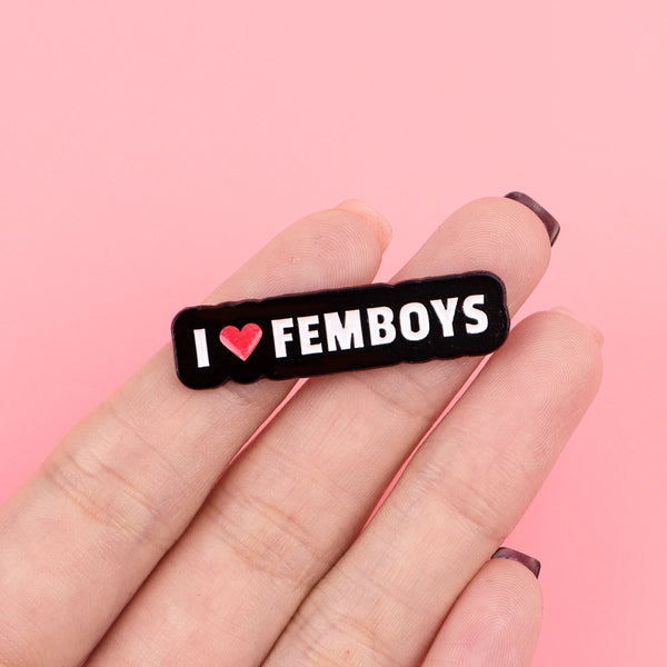 A hand holds a black enamel pin with the text I love femboys engraved on it.