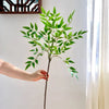 A hand holds up a tall artificial Nandina plant with bright green leaves against a white wall.
