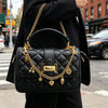 A black quilted leather handbag with a gold chain strap and multiple charms is carried by a person in an urban setting.