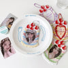 A white plate holds a strawberry-themed card holder and a bag keychain featuring strawberries and a portrait.