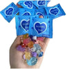 A hand holds several blue mystery bags with heart designs and a pile of glow in the dark resin decor for fairy gardens