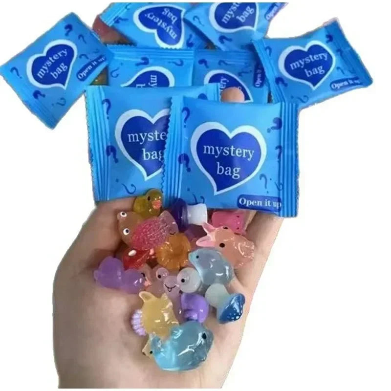 A hand holds several blue mystery bags with heart designs and a pile of glow in the dark resin decor for fairy gardens