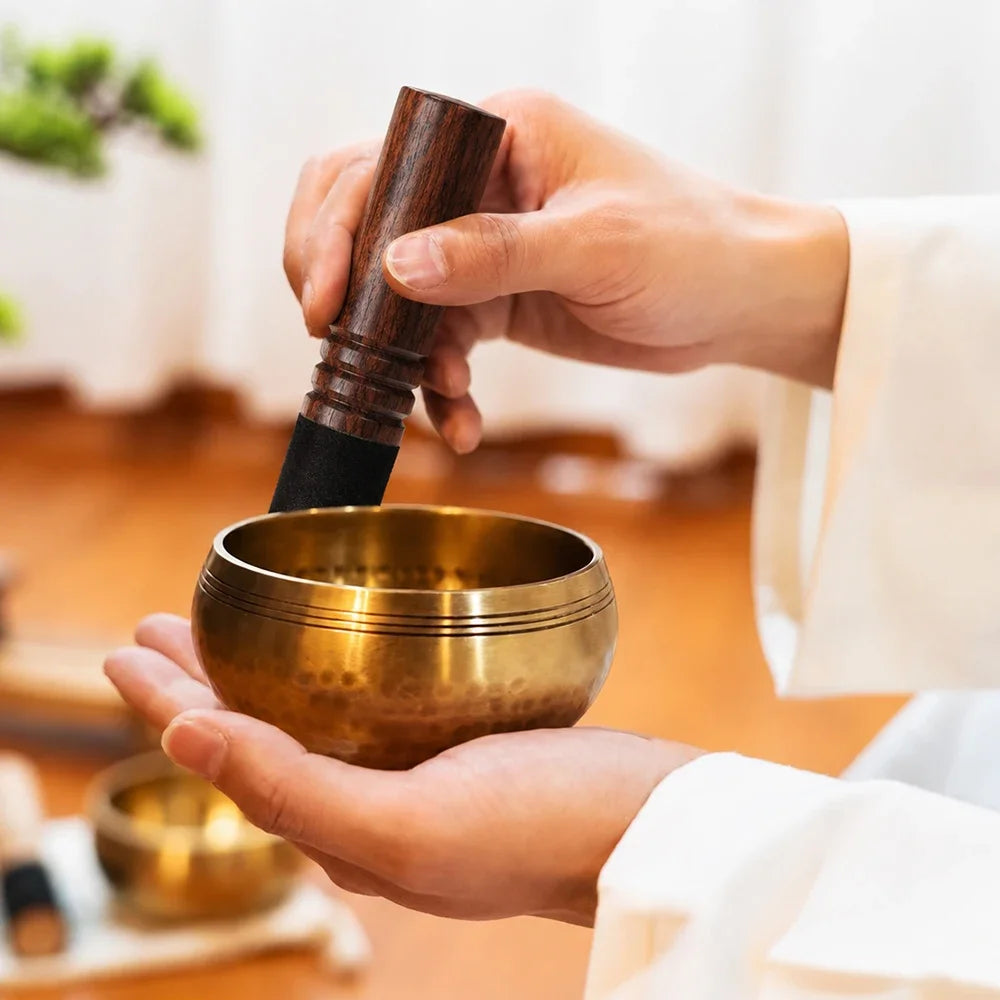 A person is holding a handcrafted copper singing bowl with a wooden mallet for meditation or sound healing.