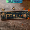 A rectangular metal wall sign with gold lettering and floral designs, featuring the text STAY WEIRD on a dark background.