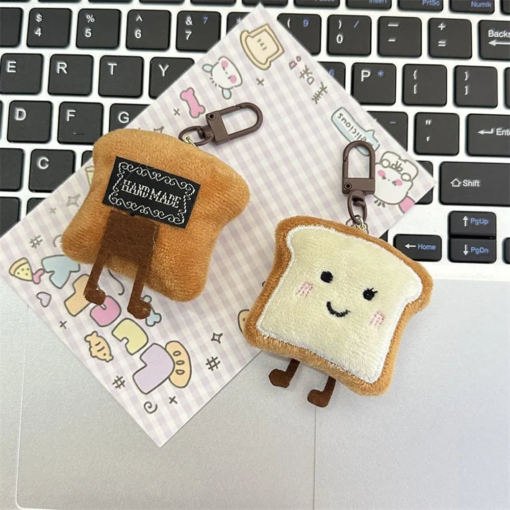A small, fluffy toast bread shaped plush charm with a keyring attached, resting on a notebook next to a laptop keyboard.