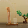 A tall glass incense holder with bamboo sticks and a metal rod is displayed on a wooden surface.