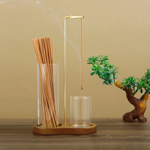 A tall glass incense holder with bamboo sticks and a metal rod is displayed on a wooden surface.