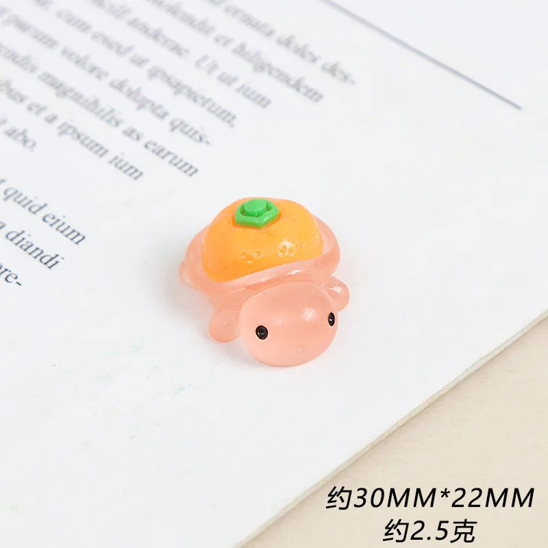 A small, pink resin charm shaped like a turtle with an orange cap and green detail resting on a white paper.