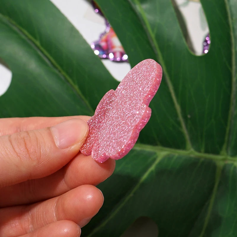 A hand holds a pink glitter acrylic charm shaped like a butterfly against a green leaf background.