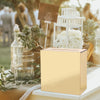 A large, rectangular gold acrylic gift box sits on a white surface surrounded by greenery and decorative elements.