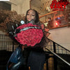A woman holds a bouquet of deep red roses with black wrapping paper in an indoor setting.
