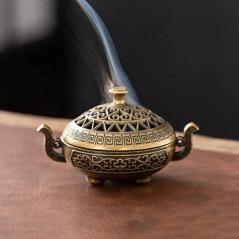 A vintage brass incense burner holder with a lid and handles, emitting smoke.