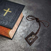 A dark leather-bound book with a gold cross on the cover next to a brown leather cord pendant necklace.