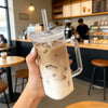 A hand holds an iced coffee drink in a clear glass mug with a handle and straw, set against a cafe background.